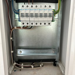 Elektryk - Wnętrze metalowej skrzynki rozdzielczej z zamontowanymi bezpiecznikami Hager MB 120A B20, widoczne kable w różnych kolorach zabezpieczone białymi opaskami zaciskowymi.