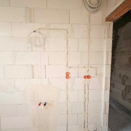 Elektryk - Ściana z betonu komórkowego w trakcie prac instalacyjnych: widoczne bruzdy pod przewody, puszki instalacyjne w kolorze pomarańczowym, peszle przy podłodze oraz zwój kabla pod sufitem.