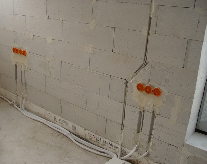 Surowa ściana z betonu komórkowego z wyciętymi bruzdami i zamontowanymi puszkami elektrycznymi, widoczne przewody w listwach przypodłogowych.