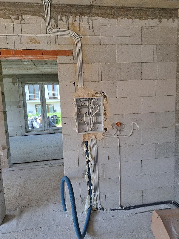 Wewnętrzna ściana z betonu komórkowego w trakcie prac elektrycznych: widoczne peszle z przewodami, puszka elektryczna z kablami, niebieski karbowany przewód na podłodze, pianka montażowa...