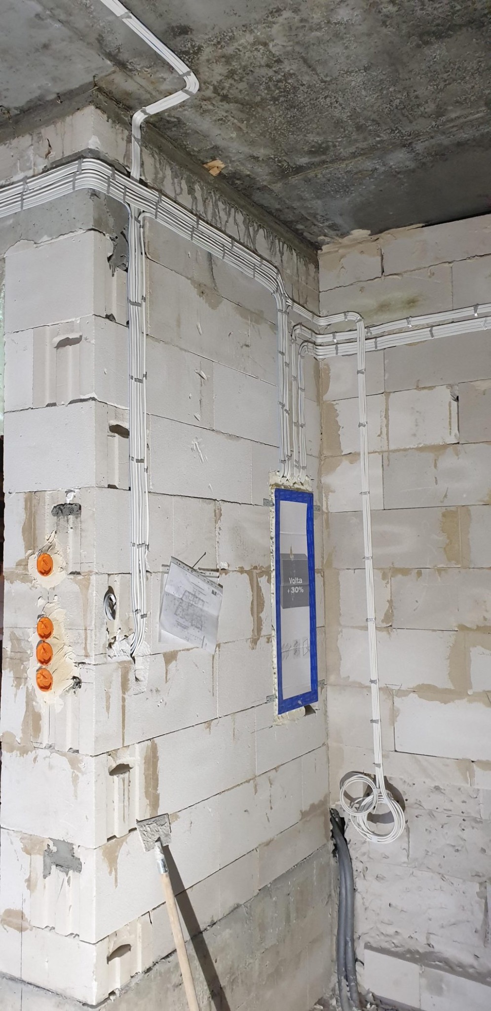 Wnętrze w trakcie prac: białe przewody elektryczne ułożone w korytkach na ścianach z bloczków, pomarańczowe puszki instalacyjne, częściowo zakryty otwór na rozdzielnicę z widocznym napisem 'Volta...