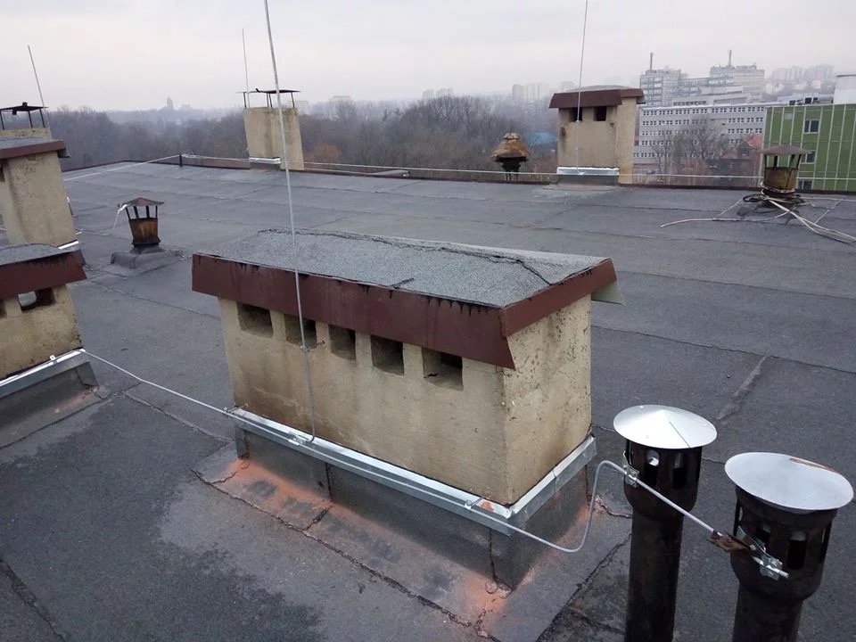 Widok na dach budynku z kominami i zamontowaną instalacją odgromową, przewody biegną wzdłuż kominów, w tle zabudowa miejska w pochmurny dzień.