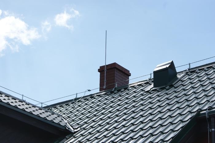Metalowy dach z zainstalowaną instalacją odgromową, widoczne przewody i maszt na kominie, błękitne niebo w tle.
