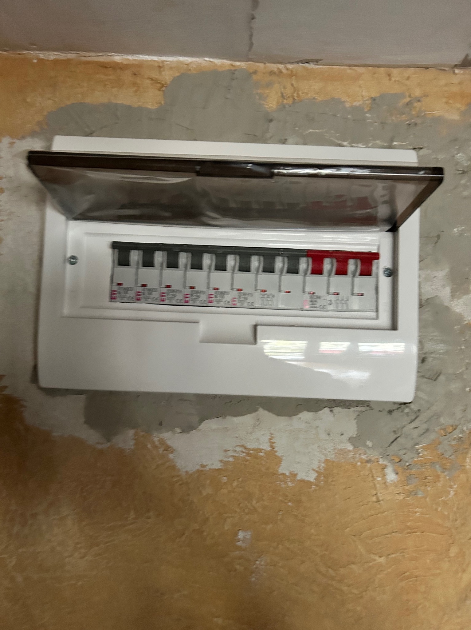 Otwarta skrzynka bezpiecznikowa, biała, z widocznymi bezpiecznikami, zamontowana na nierównej, surowej ścianie z widocznymi śladami napraw i malowania.