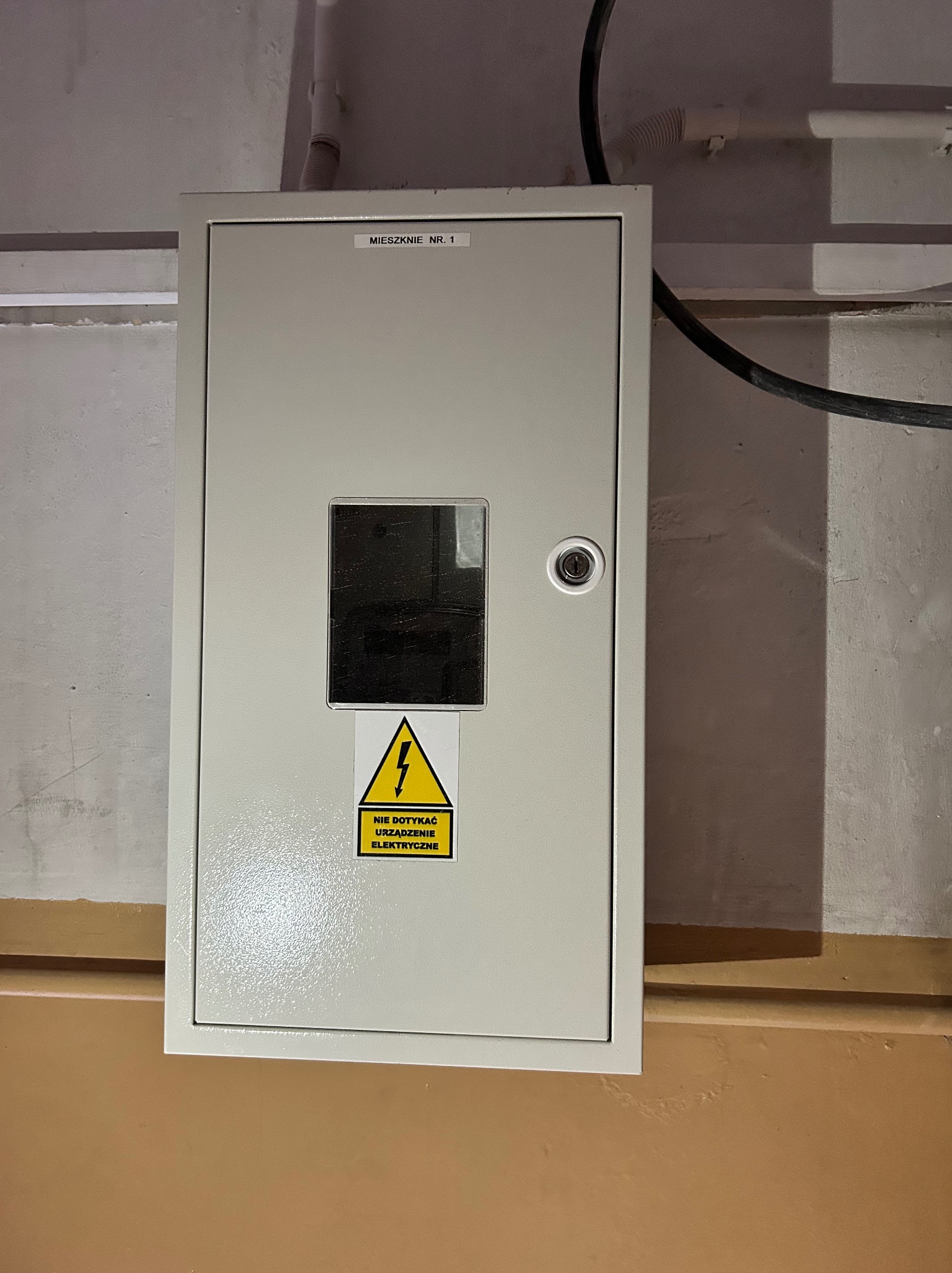 Szara skrzynka elektryczna z okienkiem i ostrzeżeniem o napięciu, oznaczona jako 'MIESZKANIE NR. 1', zamontowana na pomalowanej ścianie w piwnicy.