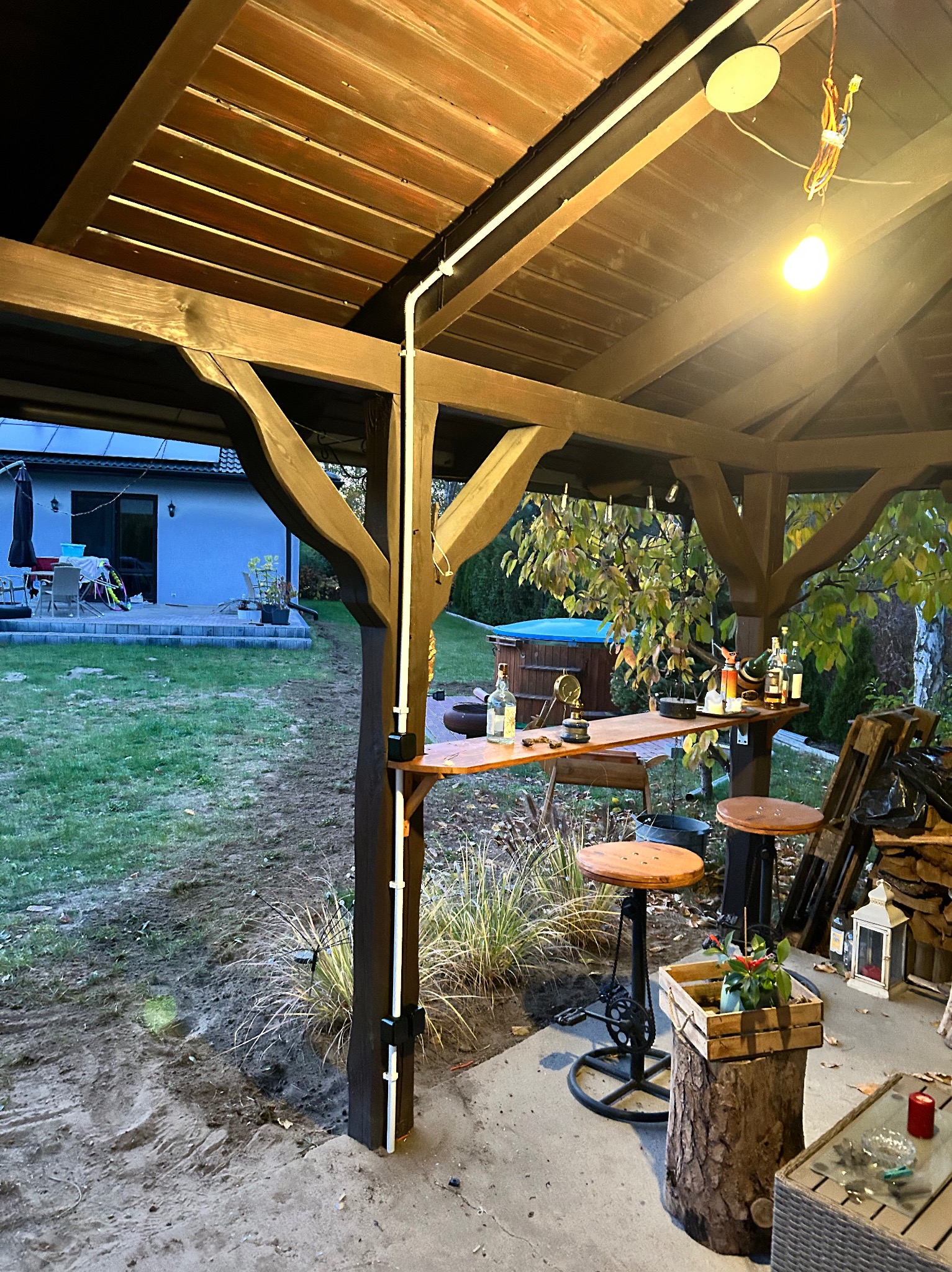 Oświetlenie LED zamontowane w drewnianej altanie z barem i stołkami barowymi, widok na ogród z jacuzzi.