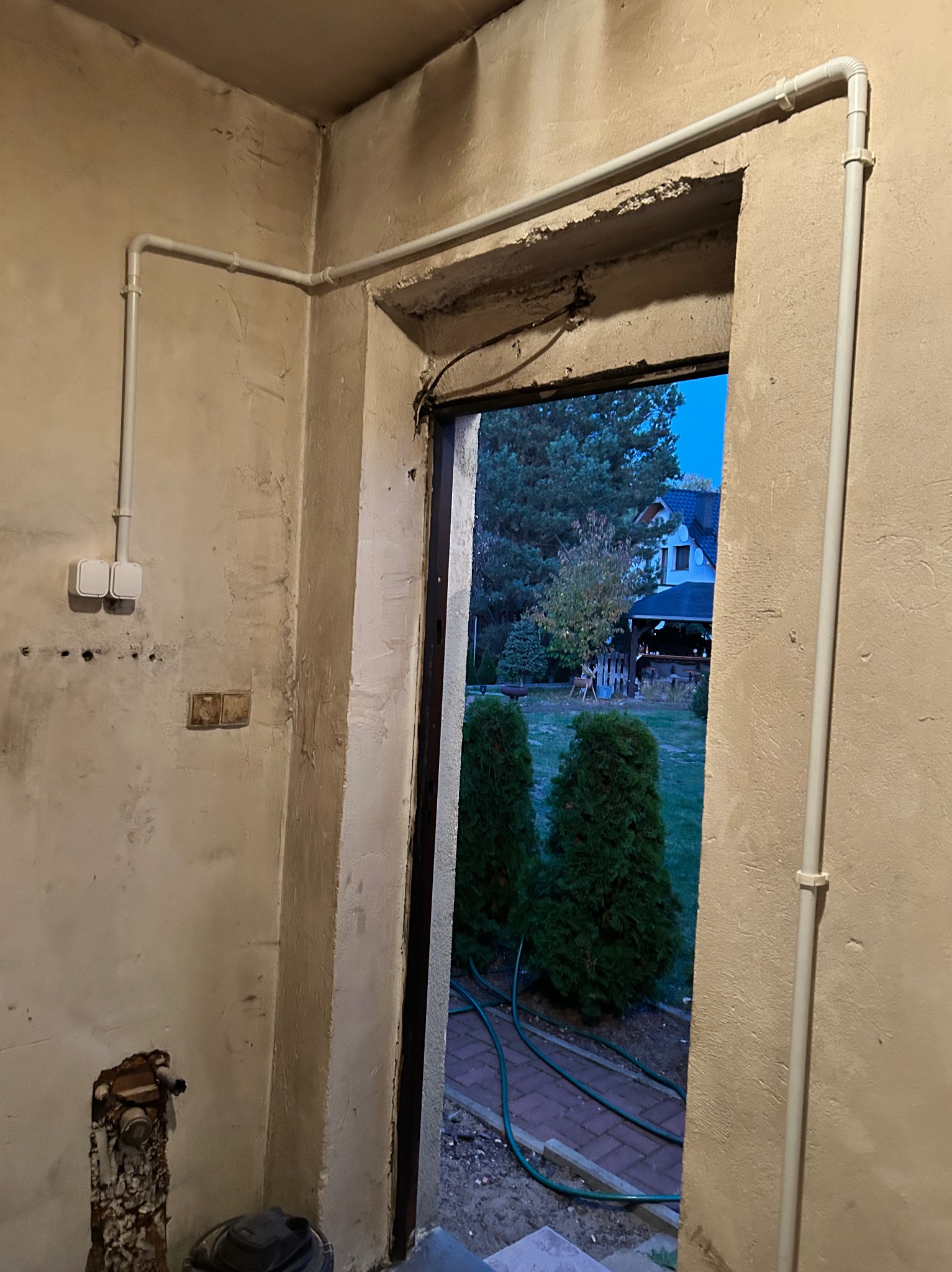 Wnętrze w trakcie remontu z widocznymi białymi rurami instalacji elektrycznej poprowadzonymi po ścianach, wystające kable i gniazdka, widok na ogród przez otwarte drzwi.