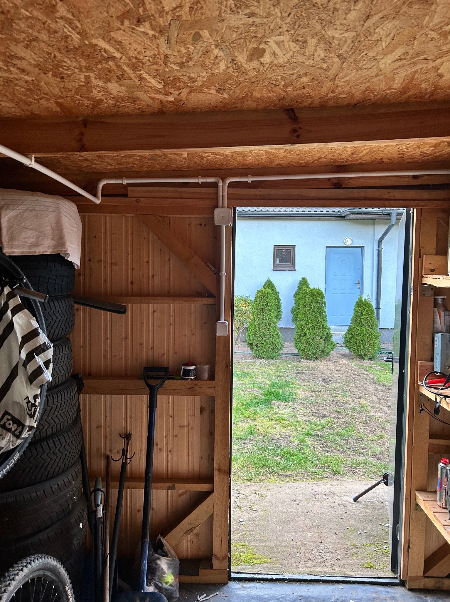 Wnętrze drewnianego garażu z widoczną instalacją elektryczną w białej rurce PCV pod sufitem, oświetleniem i stertą opon rowerowych, przez otwarte drzwi widać podwórko z trzema tujami przed domem.