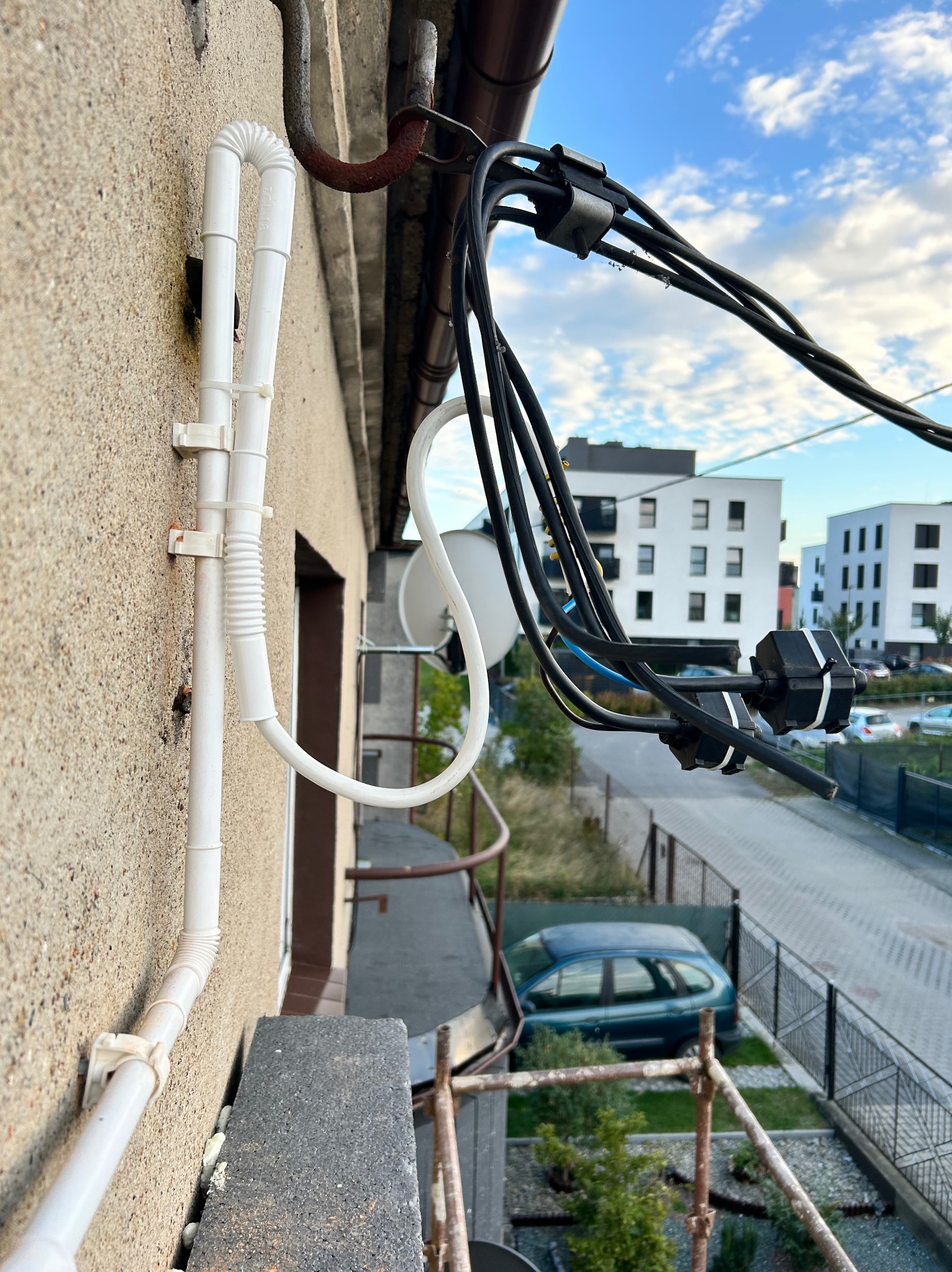 Białe rury PCV przymocowane do elewacji budynku za pomocą plastikowych uchwytów, z widoczną falistą sekcją, oraz czarne przewody elektryczne przymocowane do dachu, z widokiem na nowoczesne budynki...