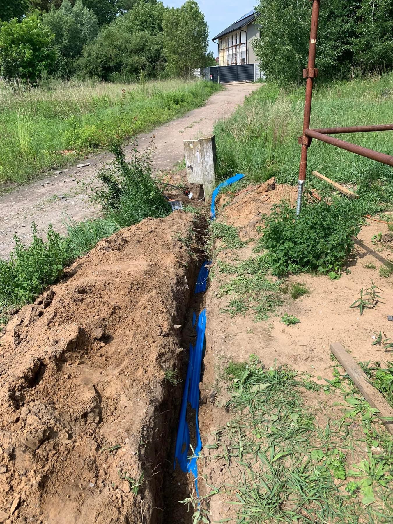 Wykop w ziemi z ułożonymi niebieskimi rurami, prawdopodobnie do przyłącza elektrycznego, widoczny betonowy słup obok wykopu oraz budynek w oddali.