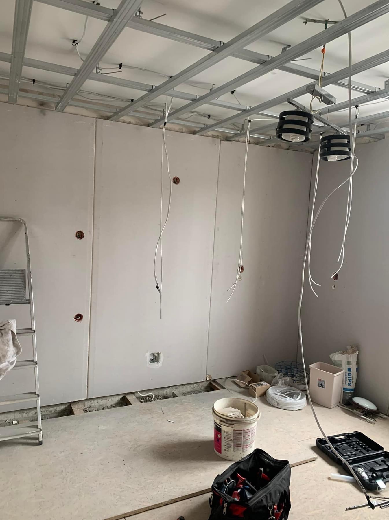 Wnętrze w trakcie remontu, widoczne przewody elektryczne wystające z sufitu podwieszanego i ścian gipsowych, drabina aluminiowa z boku, narzędzia i materiały budowlane porozrzucane na podłodze.