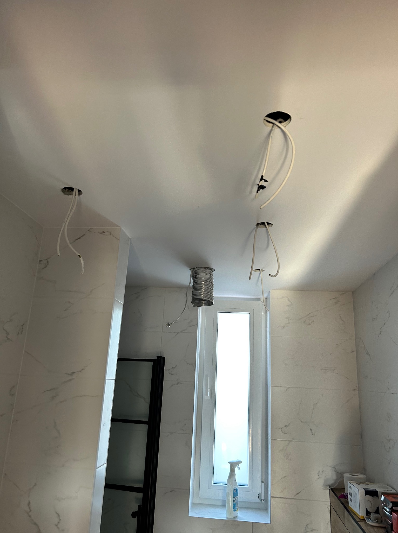 Przygotowanie do montażu oświetlenia sufitowego w łazience wykończonej płytkami imitującymi marmur. Widoczne wystające przewody z otworów w suficie oraz zwój kabla przy oknie.