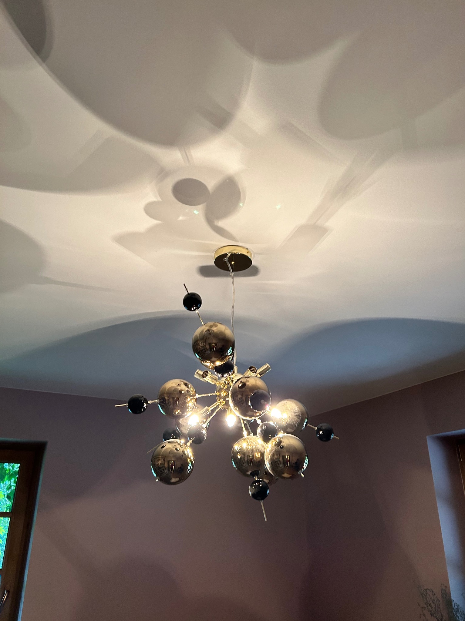 Nowoczesna lampa sufitowa z kulistymi kloszami w odcieniach złota i czerni, rzucająca dekoracyjne cienie na jasny sufit w pomieszczeniu z oknami.