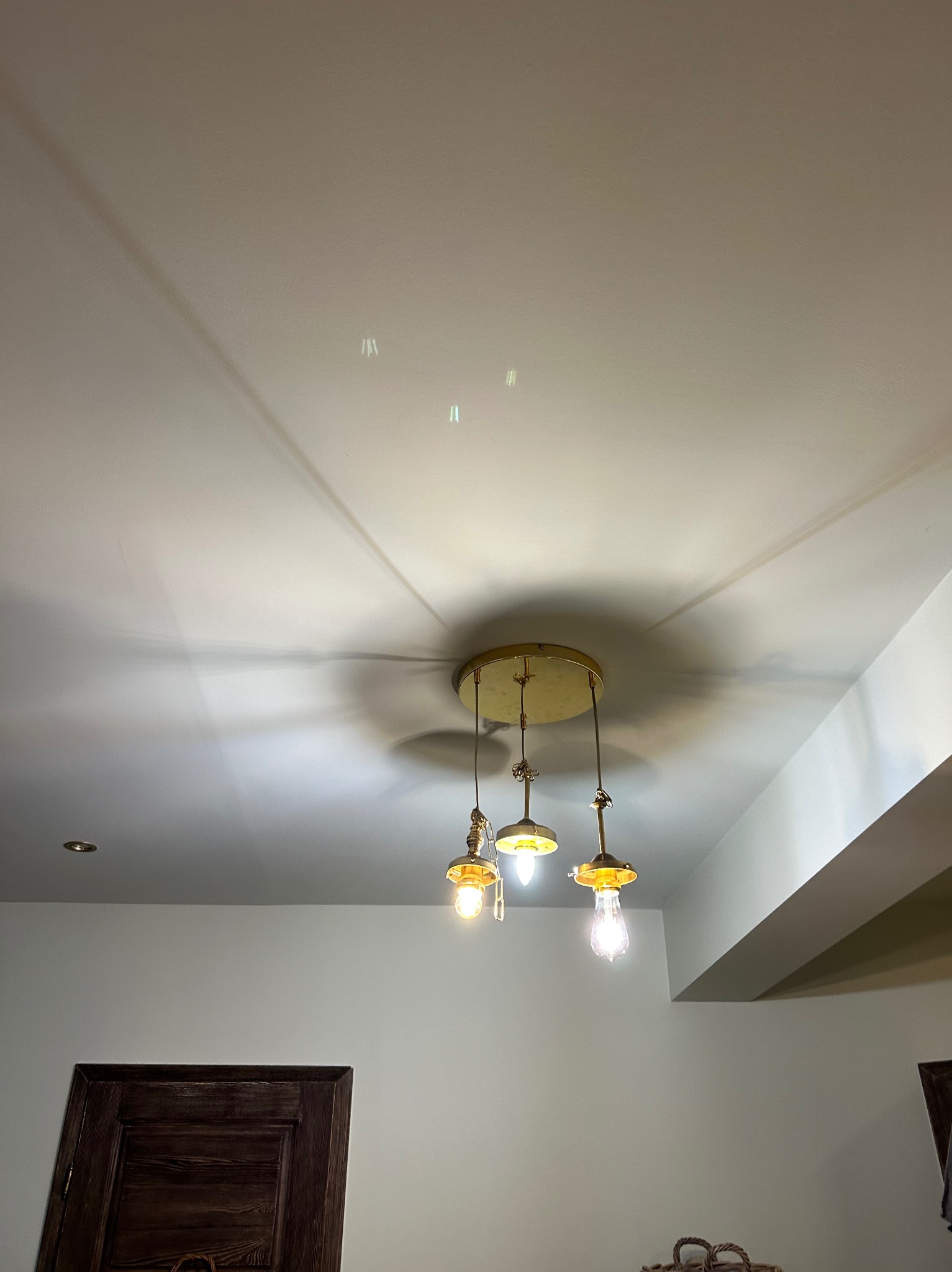 Złota lampa wisząca z trzema żarówkami w stylu vintage, rzucająca cienie na biały sufit w jasnym wnętrzu.