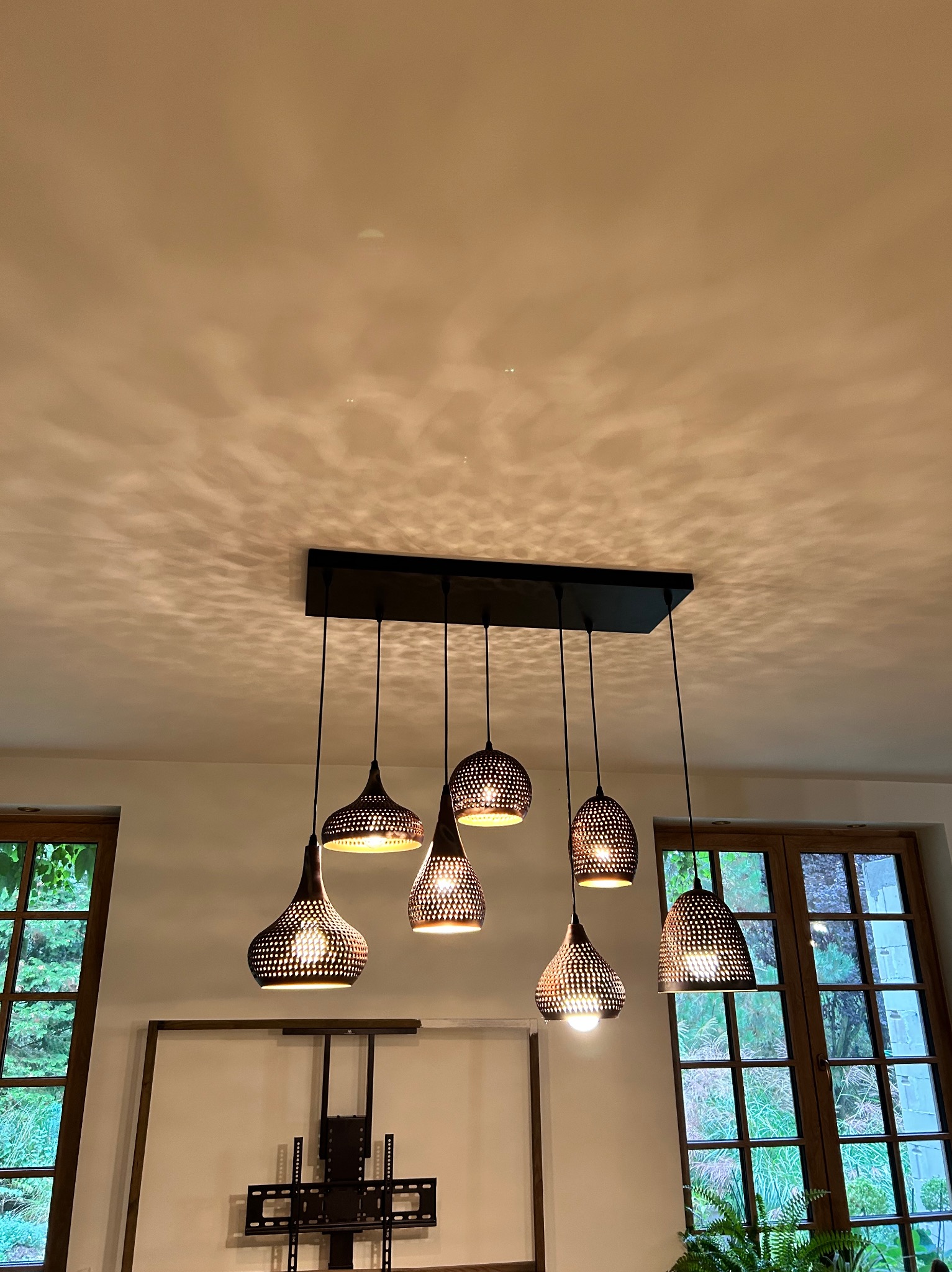 Nowoczesna lampa wisząca z sześcioma ażurowymi kloszami, zawieszona na prostokątnej listwie, rzucająca dekoracyjne cienie na sufit w ciepłym odcieniu.