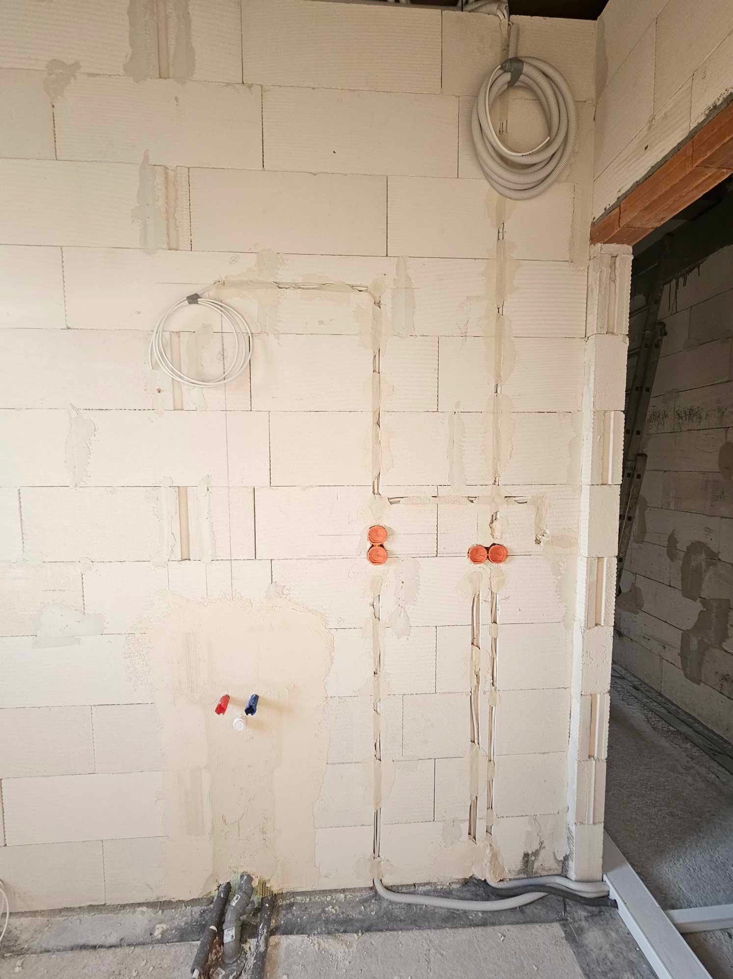 Ściana z betonu komórkowego w trakcie prac instalacyjnych: widoczne bruzdy pod przewody, puszki instalacyjne w kolorze pomarańczowym, peszle przy podłodze oraz zwój kabla pod sufitem.