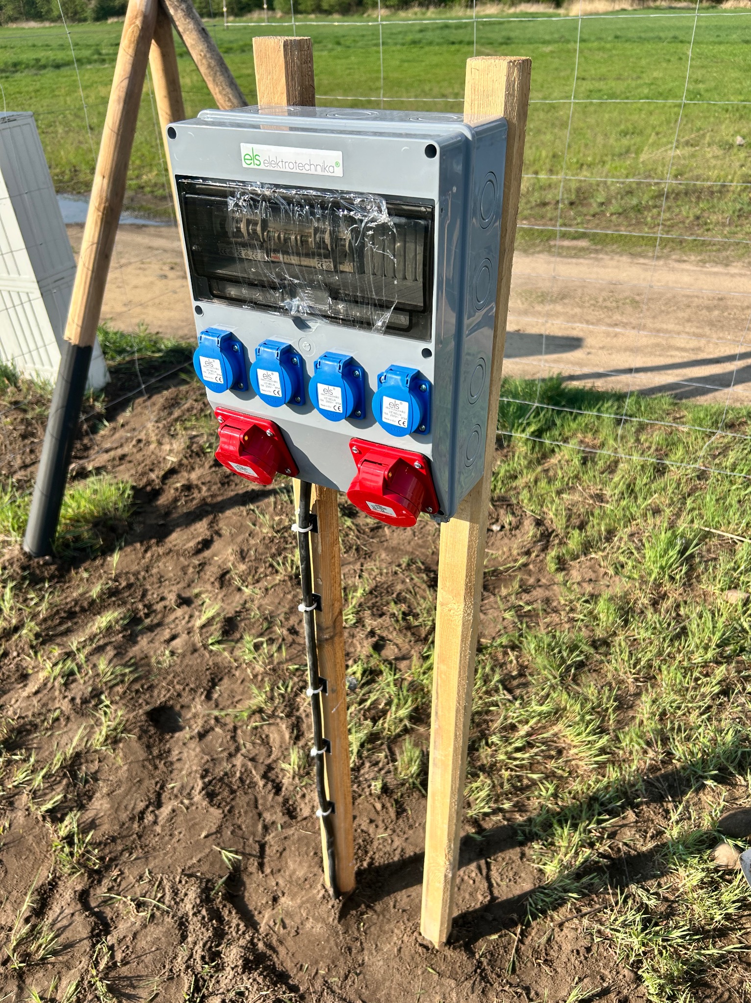 Szara skrzynka elektryczna z niebieskimi i czerwonymi gniazdami, zamontowana na drewnianych słupkach na zewnątrz, z widocznymi przewodami i zielonym tłem.