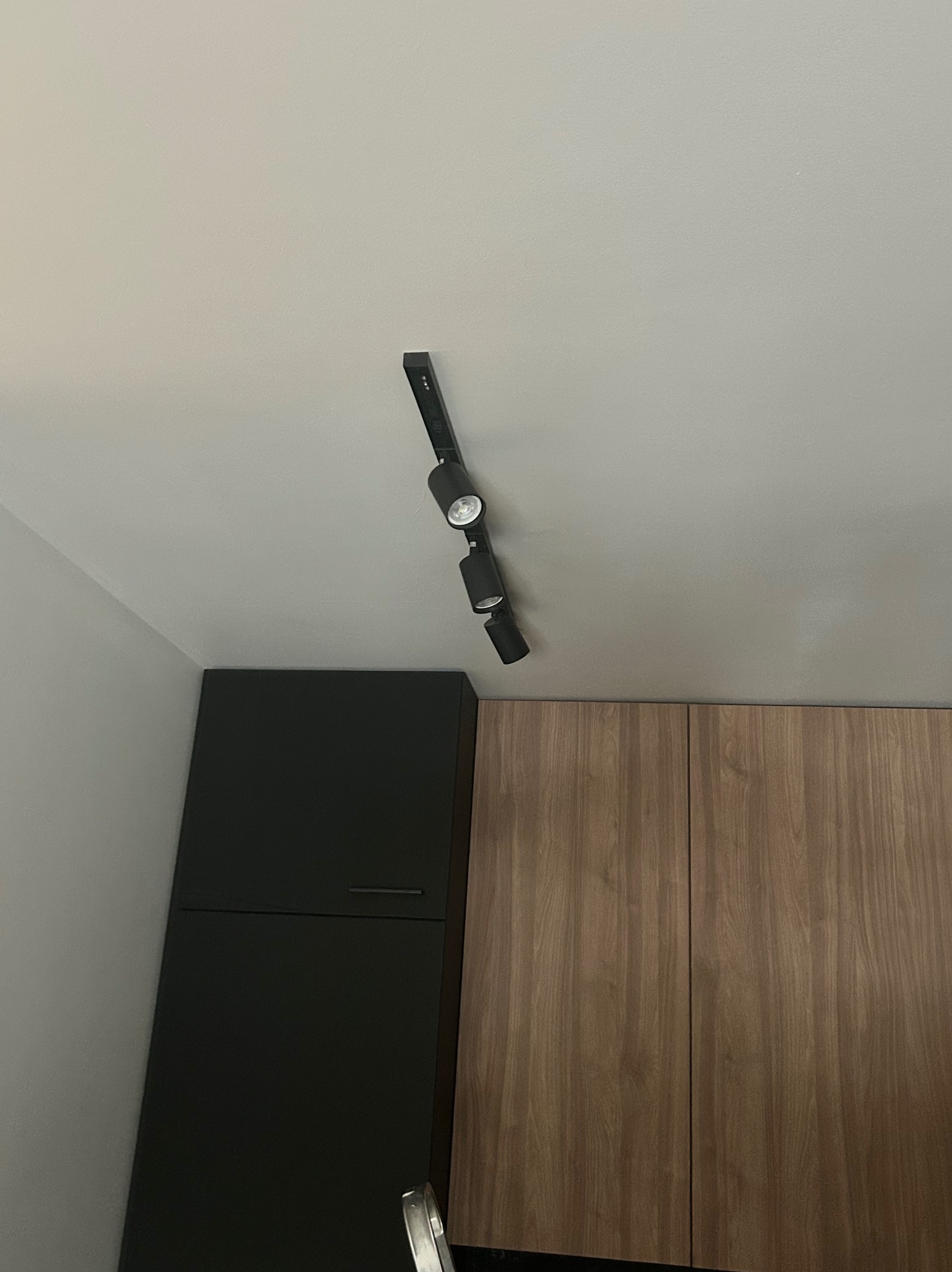 Czarna listwa z trzema reflektorami LED zamontowana na suficie w kuchni, oświetlająca szafki w kolorze drewna i czarne szafki. Minimalistyczny design.