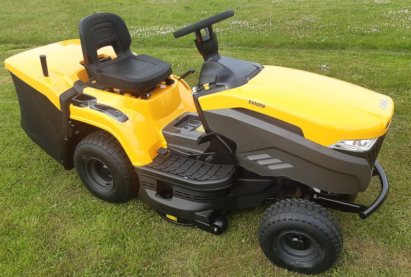 Żółta kosiarka samojezdna marki Stiga, model Estate, stojąca na trawniku z widocznymi drobnymi kwiatami w tle.