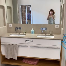 Tedeco - Nowoczesna łazienka z podwójną umywalką na blacie z jasnego kamienia, białe szafki z poziomymi uchwytami, drewniana półka z ręcznikami, duże lustro z odbiciem osoby robiącej zdjęcie telefonem.