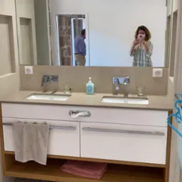 Nowoczesna łazienka z podwójną umywalką na blacie z jasnego kamienia, białe szafki z poziomymi uchwytami, drewniana półka z ręcznikami, duże lustro z odbiciem osoby robiącej zdjęcie telefonem.