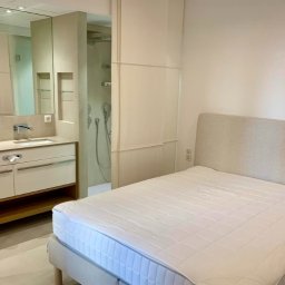 Tedeco - Nowoczesna sypialnia z łazienką en-suite w neutralnej kolorystyce. Widoczna umywalka z dwoma kranami, kabina prysznicowa i łóżko z beżowym zagłówkiem.