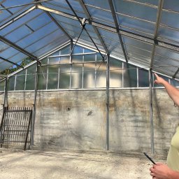 Tedeco - Wnętrze szklarni z betonową ścianą i metalową konstrukcją dachu. Mężczyzna w okularach wskazuje na górę, trzymając tablet. Widoczne ślady użytkowania i potencjalne prace remontowe.