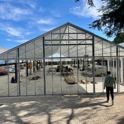 Tedeco - Konstrukcja szklarni z metalowym szkieletem w trakcie budowy, widok z zewnątrz, mężczyzna wchodzi do środka. W tle błękitne niebo i częściowo widoczne inne budynki.