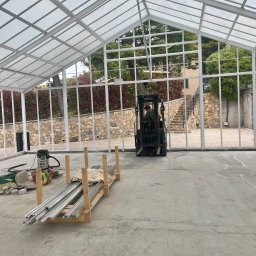 Tedeco - Montaż konstrukcji szklarni z użyciem wózka widłowego. Widoczne profile aluminiowe na palecie, odkurzacz przemysłowy i elementy konstrukcyjne.