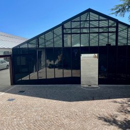 Tedeco - Przeszklona konstrukcja w Wrocławiu, prawdopodobnie szklarnia po modyfikacji, z ciemnymi szybami i metalową ramą, widok z zewnątrz na brukowany plac.