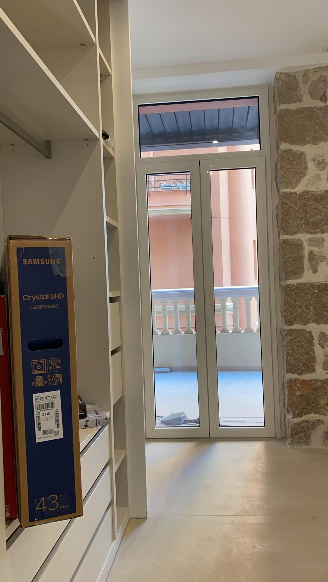 Wnętrze garderoby z białymi szafami wnękowymi, karton po telewizorze Samsung Crystal UHD 43 cali oparty o półkę, widok na balkon przez białe, dwuskrzydłowe drzwi balkonowe, ściana z kamienia...