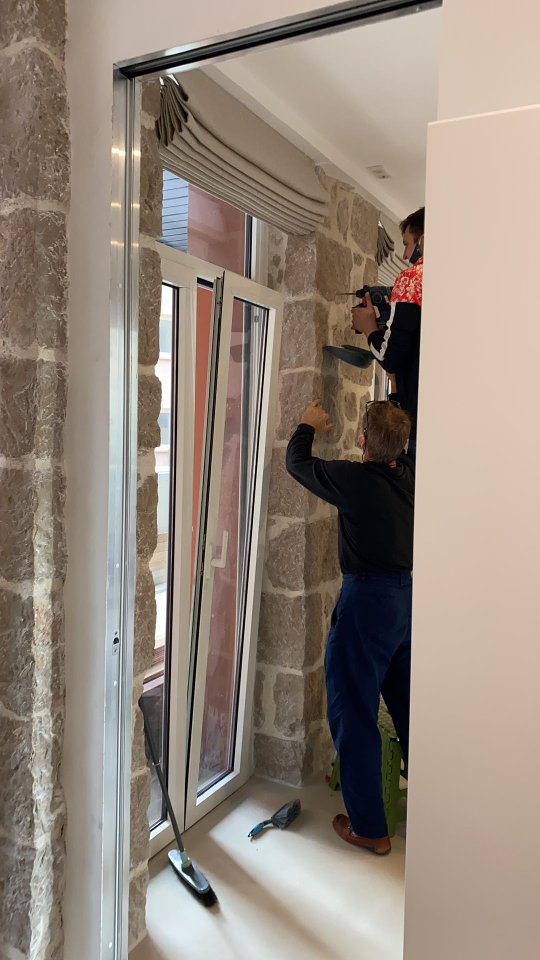 Dwóch monterów instaluje białe okno PCV w kamiennej ścianie, widoczne narzędzia i drabina.