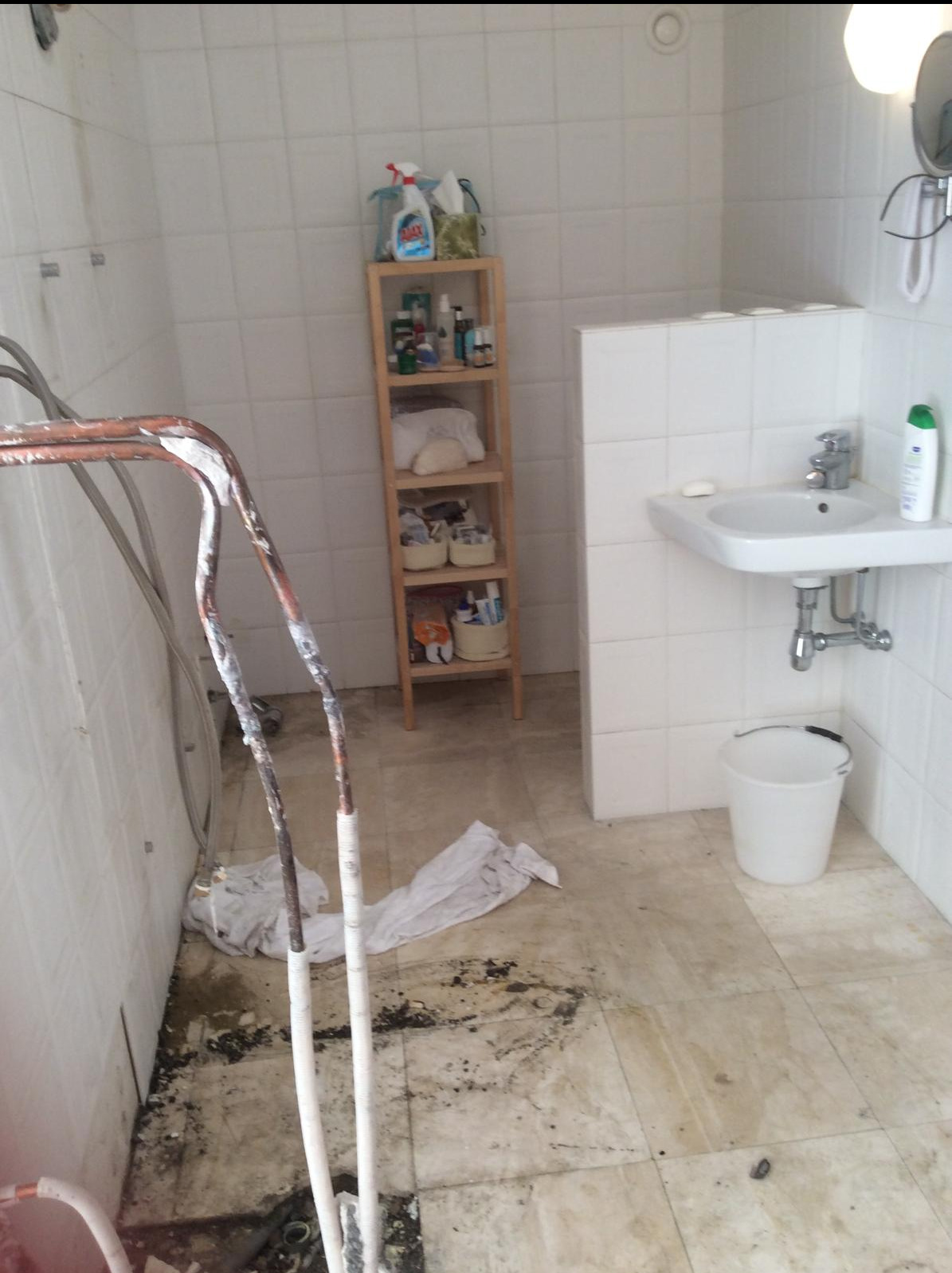 Łazienka w trakcie remontu: widoczne odsłonięte rury z resztkami starej instalacji, białe płytki na ścianach, prowizoryczna półka z kosmetykami i środkami czystości, oraz białe wiadro pod umywalką.