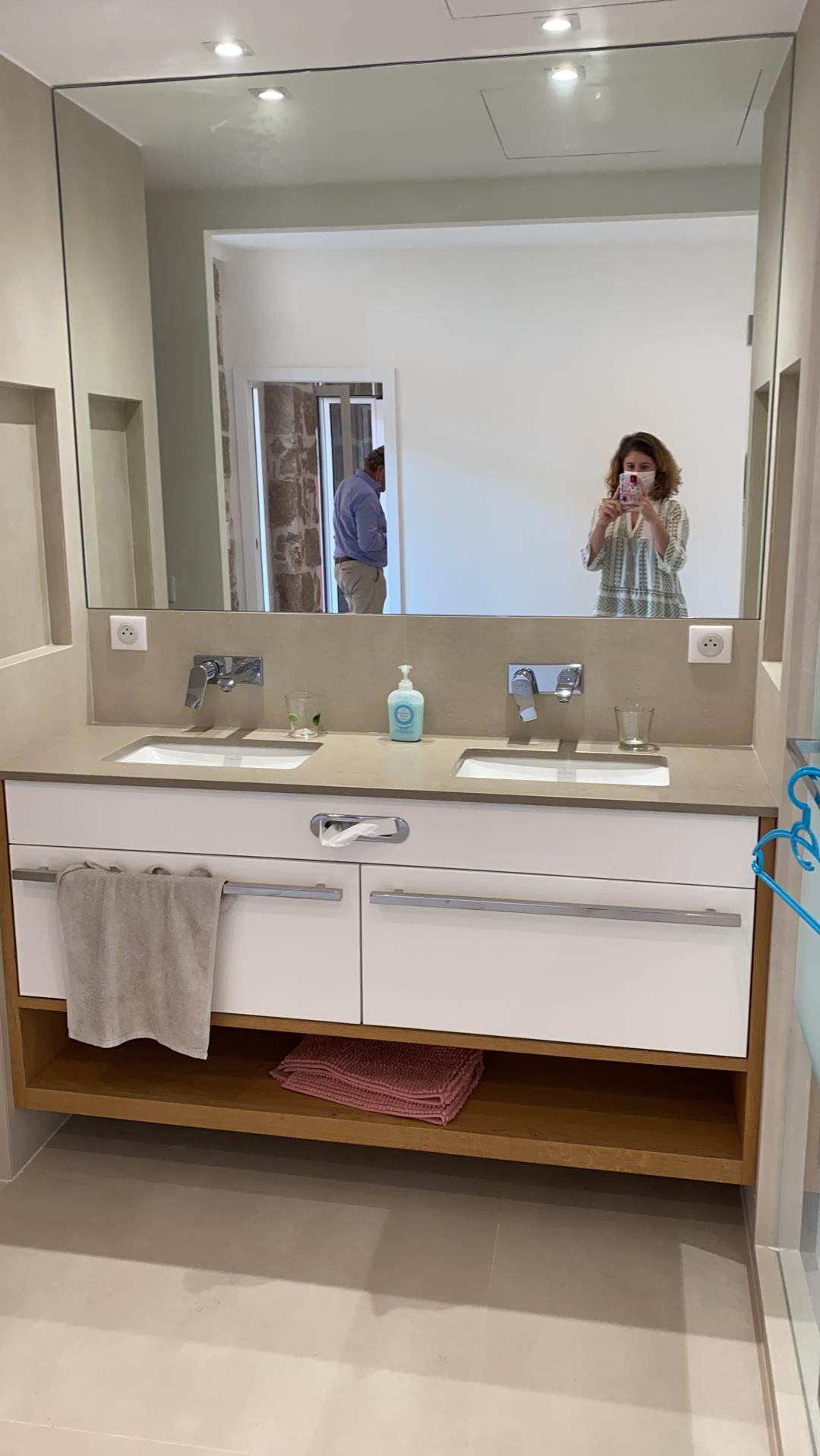 Nowoczesna łazienka z podwójną umywalką na blacie z jasnego kamienia, białe szafki z poziomymi uchwytami, drewniana półka z ręcznikami, duże lustro z odbiciem osoby robiącej zdjęcie telefonem.