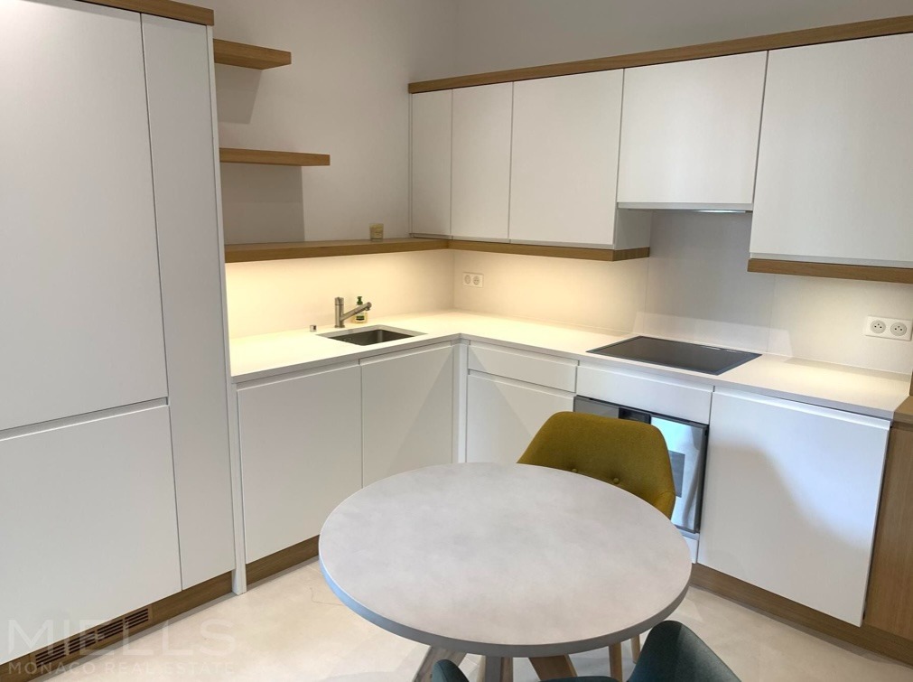 Nowoczesna, jasna kuchnia z białymi szafkami i drewnianymi akcentami. Okrągły stół z krzesłami na pierwszym planie. Minimalistyczny design, oświetlenie LED pod szafkami.