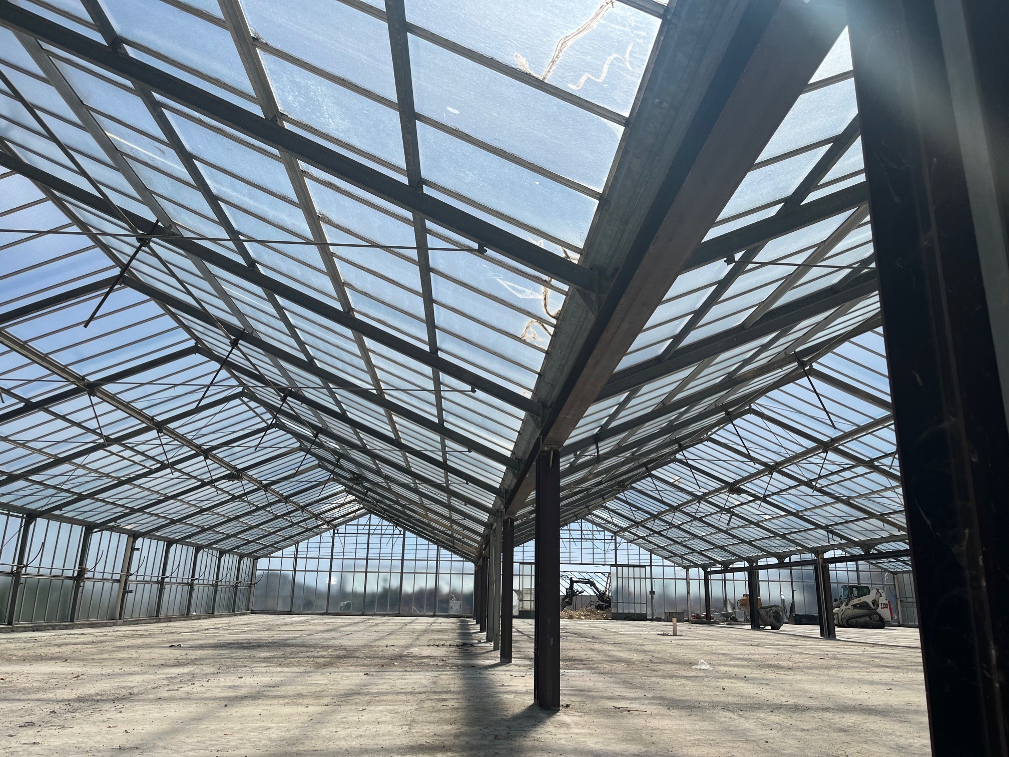 Wnętrze szklarni z metalową konstrukcją i szklanym dachem, widok na niebo. W tle widoczne maszyny budowlane, sugerujące prace remontowe lub budowlane.