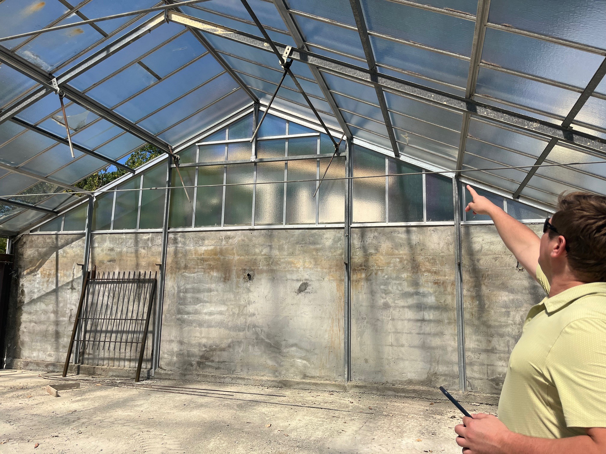 Wnętrze szklarni z betonową ścianą i metalową konstrukcją dachu. Mężczyzna w okularach wskazuje na górę, trzymając tablet. Widoczne ślady użytkowania i potencjalne prace remontowe.