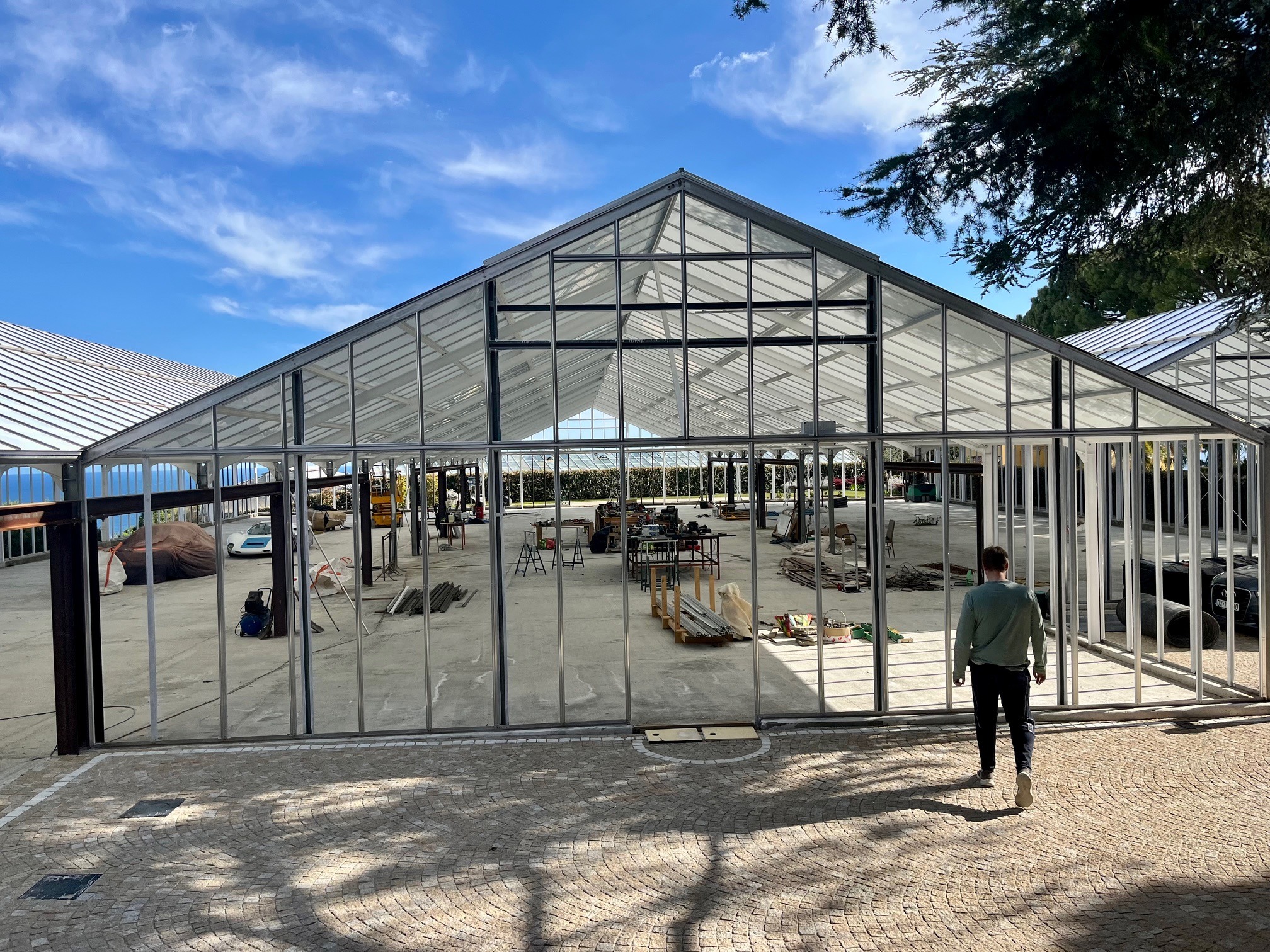 Konstrukcja szklarni z metalowym szkieletem w trakcie budowy, widok z zewnątrz, mężczyzna wchodzi do środka. W tle błękitne niebo i częściowo widoczne inne budynki.