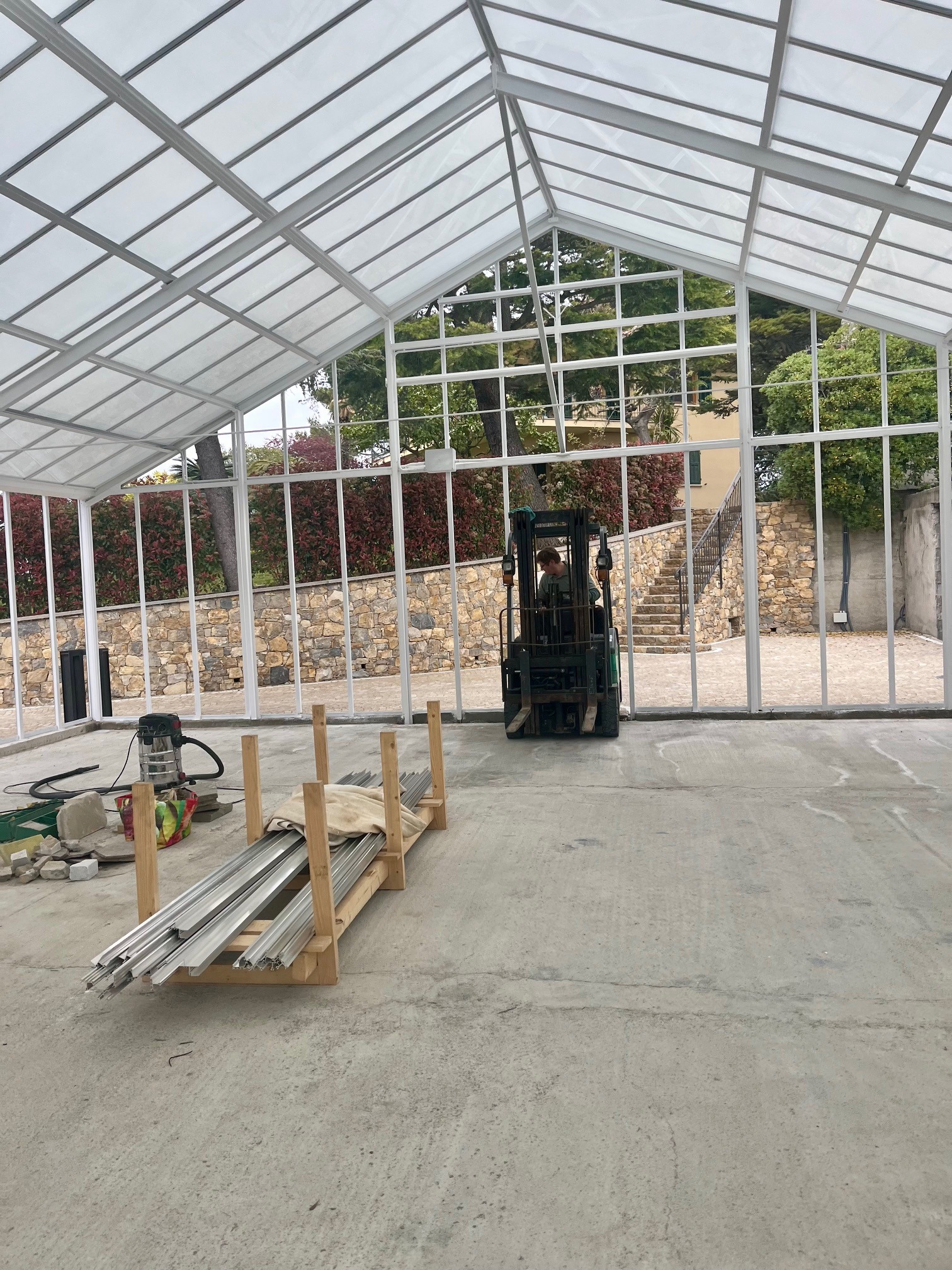 Montaż konstrukcji szklarni z użyciem wózka widłowego. Widoczne profile aluminiowe na palecie, odkurzacz przemysłowy i elementy konstrukcyjne.