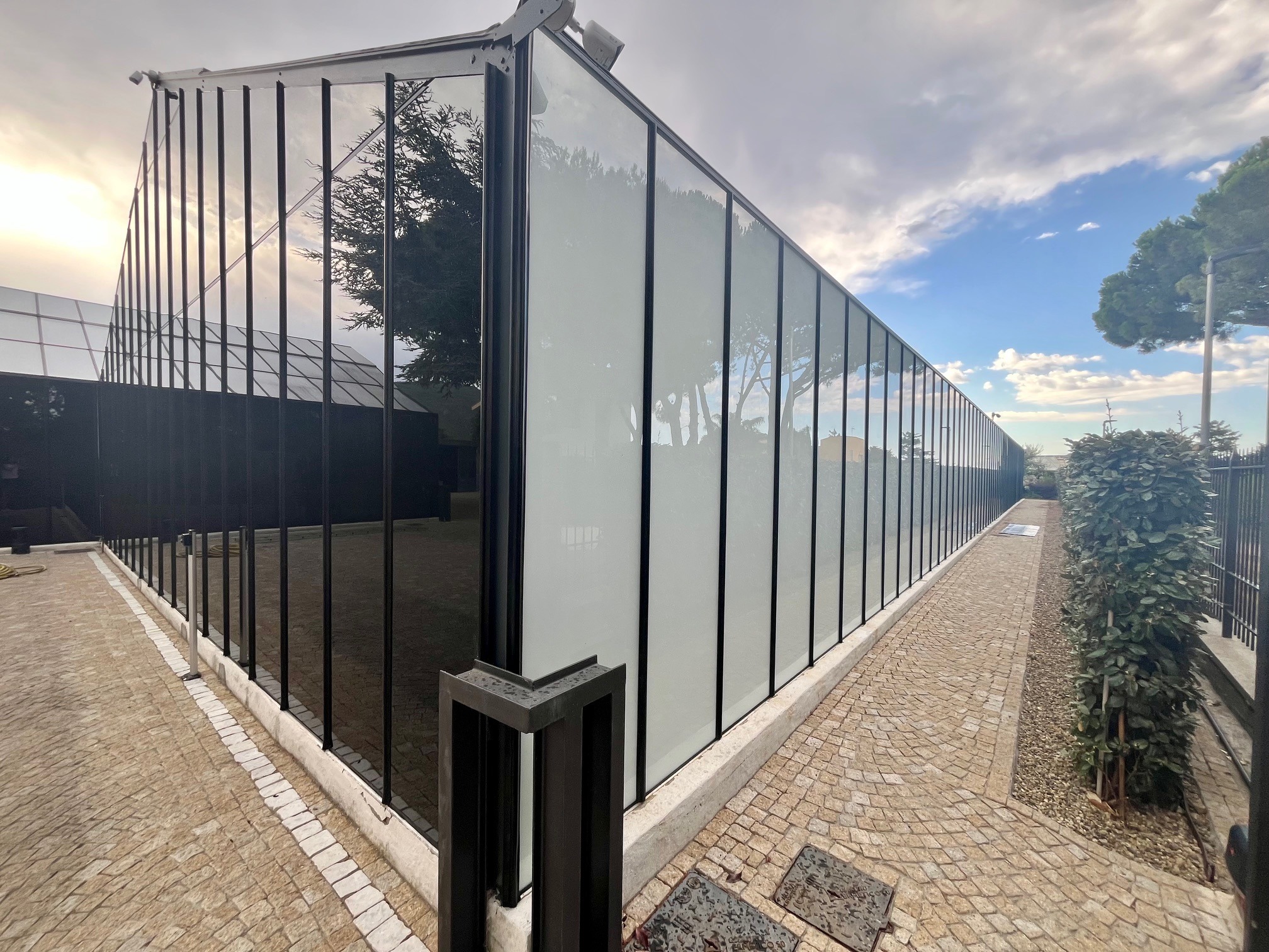 Szklana fasada budynku z czarnymi ramami, widok z zewnątrz na brukowany chodnik i zieleń. Część okien jest zasłonięta białymi roletami.