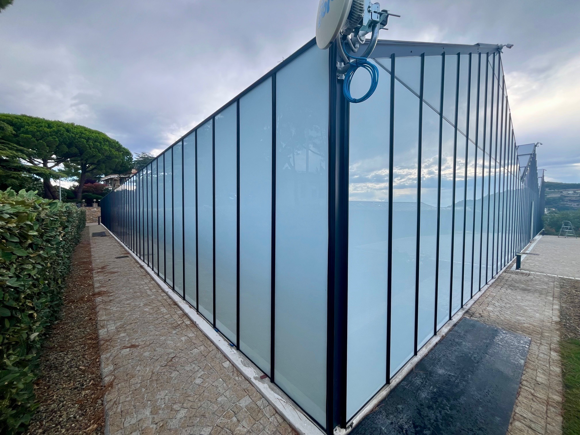 Przemiana szklarni w showroom: przeszklona konstrukcja z czarnymi ramami, widok z zewnątrz, kostka brukowa i zieleń w tle, pochmurne niebo.