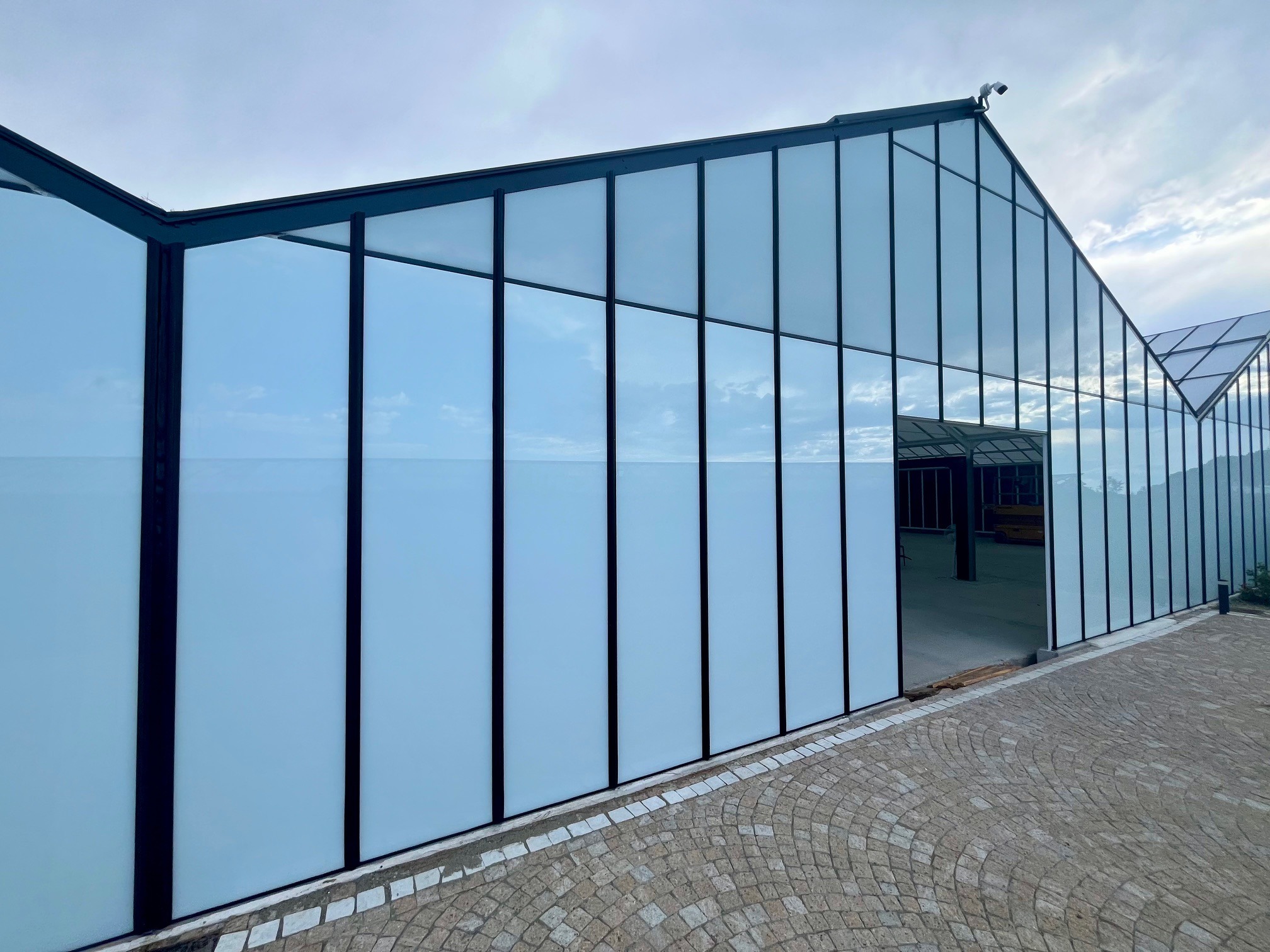 Przemiana szklarni w showroom: przeszklona konstrukcja z czarnymi ramami, widok z zewnątrz na kamienny chodnik i niebo odbijające się w szybach. Modernizacja obiektu.