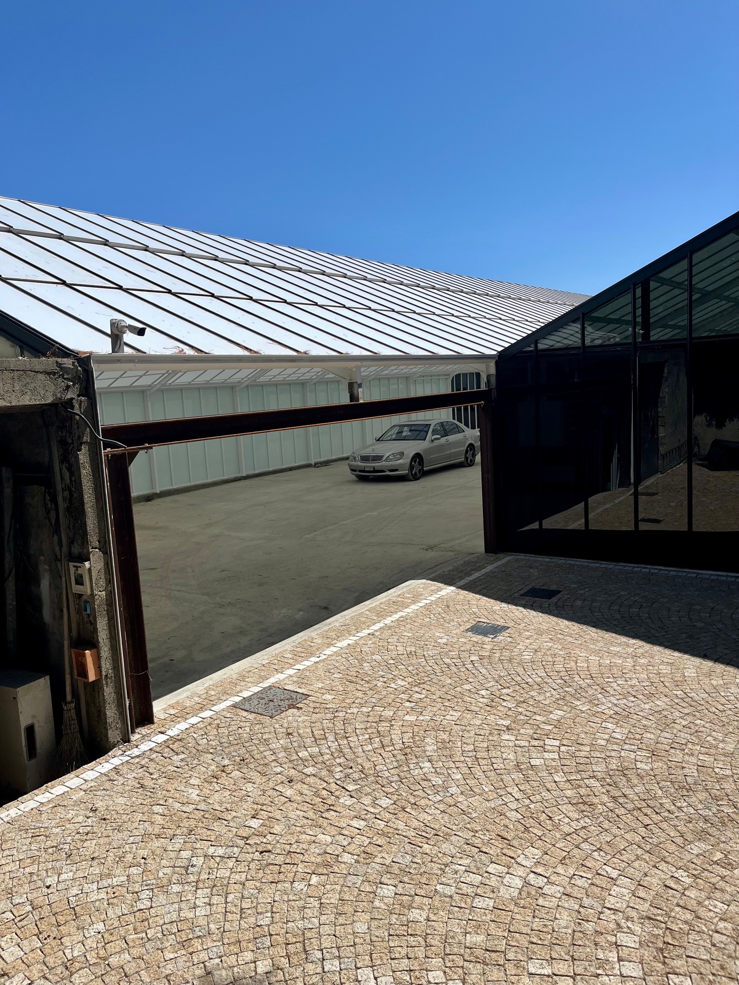 Widok z góry na dziedziniec z kostki brukowej, nowoczesny budynek z przeszkloną fasadą i srebrny samochód w tle, oświetlone jasnym, naturalnym światłem.