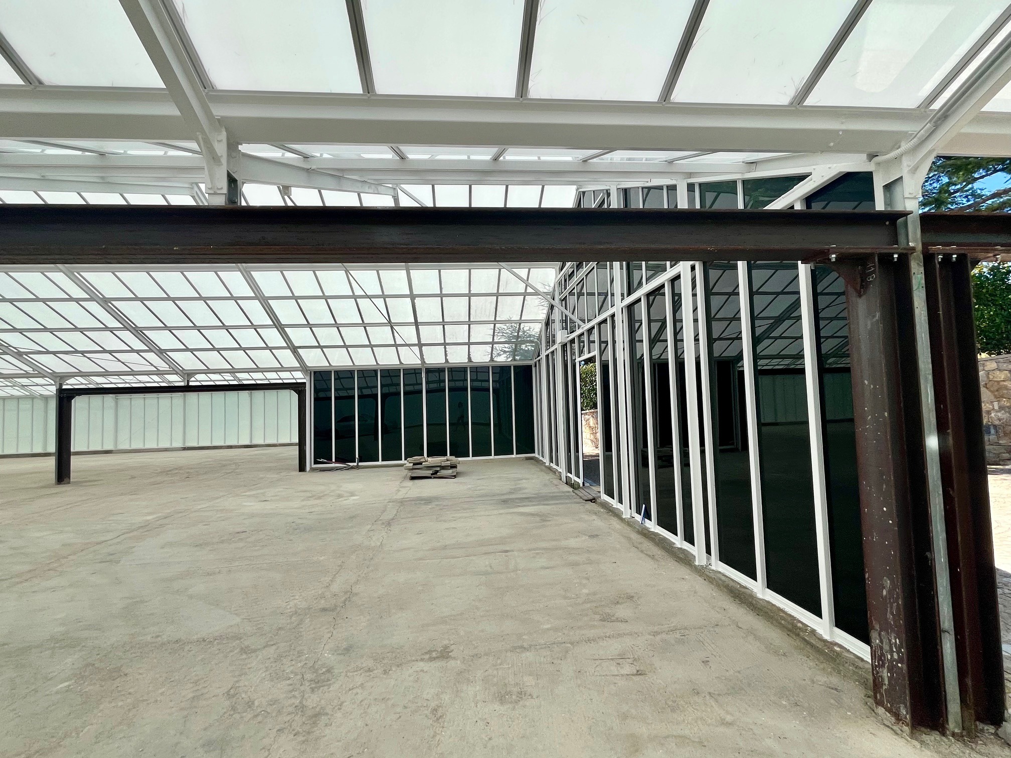 Przestronne wnętrze hali z przeszklonym dachem i ścianami. Stalowa konstrukcja pomalowana na biało. Betonowa posadzka. Widoczne elementy konstrukcyjne i fragmenty szyb.