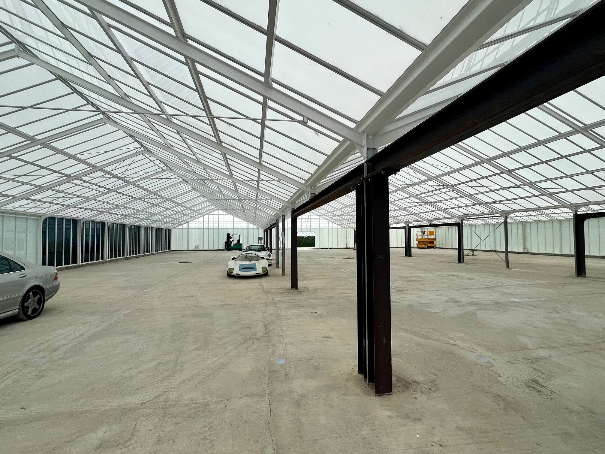 Przestronne wnętrze hali z metalową konstrukcją dachu i betonową podłogą, przygotowane na showroom samochodowy. Widoczne auta i podnośnik koszowy w tle.