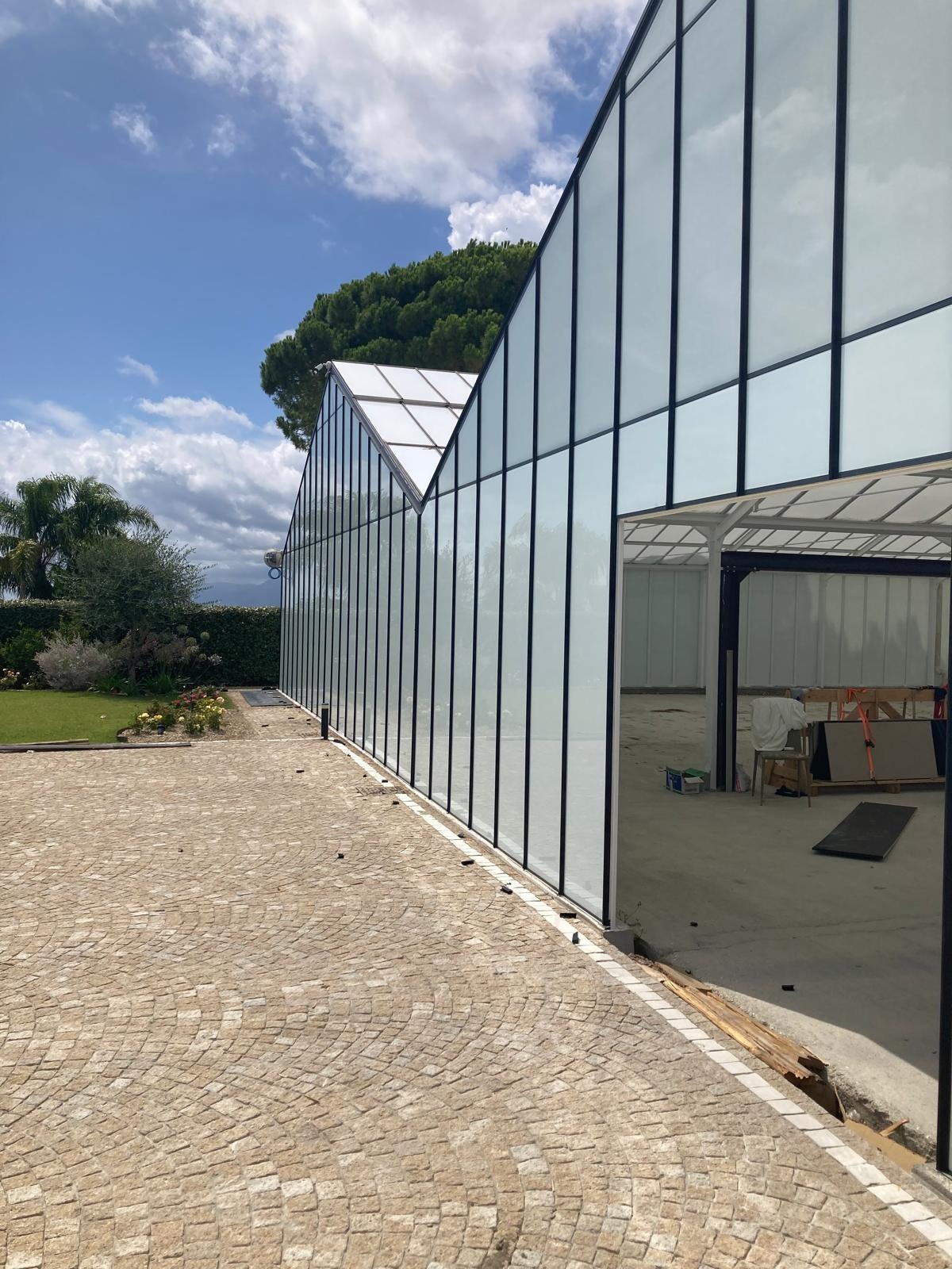 Szklana konstrukcja budynku z czarnymi ramami, widok z zewnątrz na brukowany chodnik i zarys zieleni. Wnętrze budynku w trakcie prac wykończeniowych.