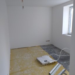 Przemysław Jóźwiak - Pokój w trakcie przygotowania do malowania: zabezpieczona folią podłoga, widoczne narzędzia malarskie, gładkie, białe ściany, surowe gniazdka elektryczne, okno z widocznymi uszczelkami.