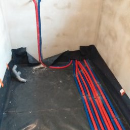 Przemysław Jóźwiak - Surowy stan łazienki w trakcie remontu: widoczne rury instalacji wodnej (czerwone i niebieskie) wychodzące ze ściany i rozłożone na czarnej folii hydroizolacyjnej, dodatkowo fragment kanalizacji...