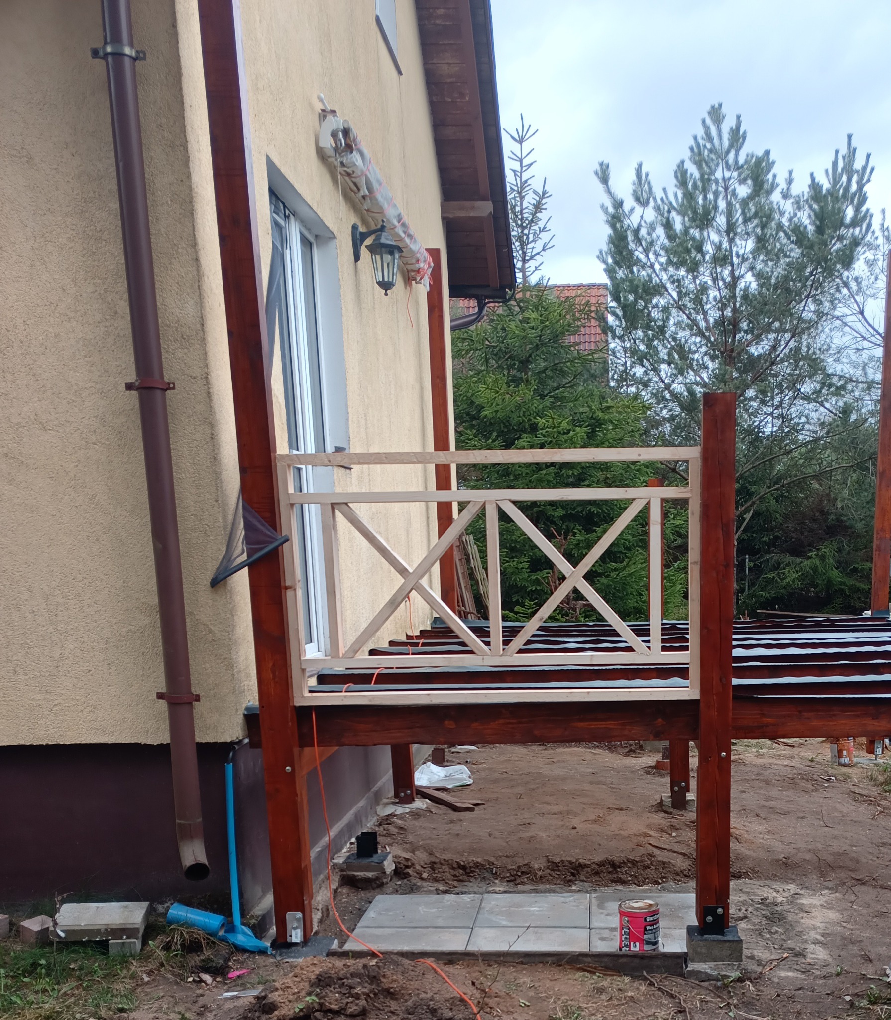 W trakcie budowy drewnianego tarasu przy domu z beżową elewacją, widoczne elementy konstrukcyjne z surowego drewna i pomalowane na brązowo, na ziemi leżą betonowe płyty i puszka farby.