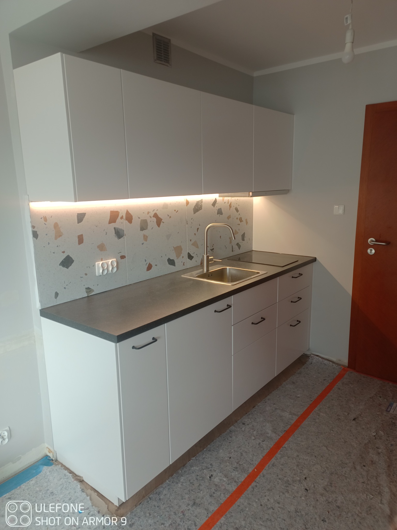 Nowoczesna kuchnia z białymi szafkami, szarym blatem i mozaikową ścianą nad zlewem, podświetlona listwą LED, widoczny fragment płyty indukcyjnej i niedokończona podłoga z taśmą zabezpieczającą.